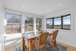 A dining area with a glass-top table, wicker chairs, and large windows offering views of the outdoors. - Oceanfront Stay on Plum Island Sound (Ipswich)