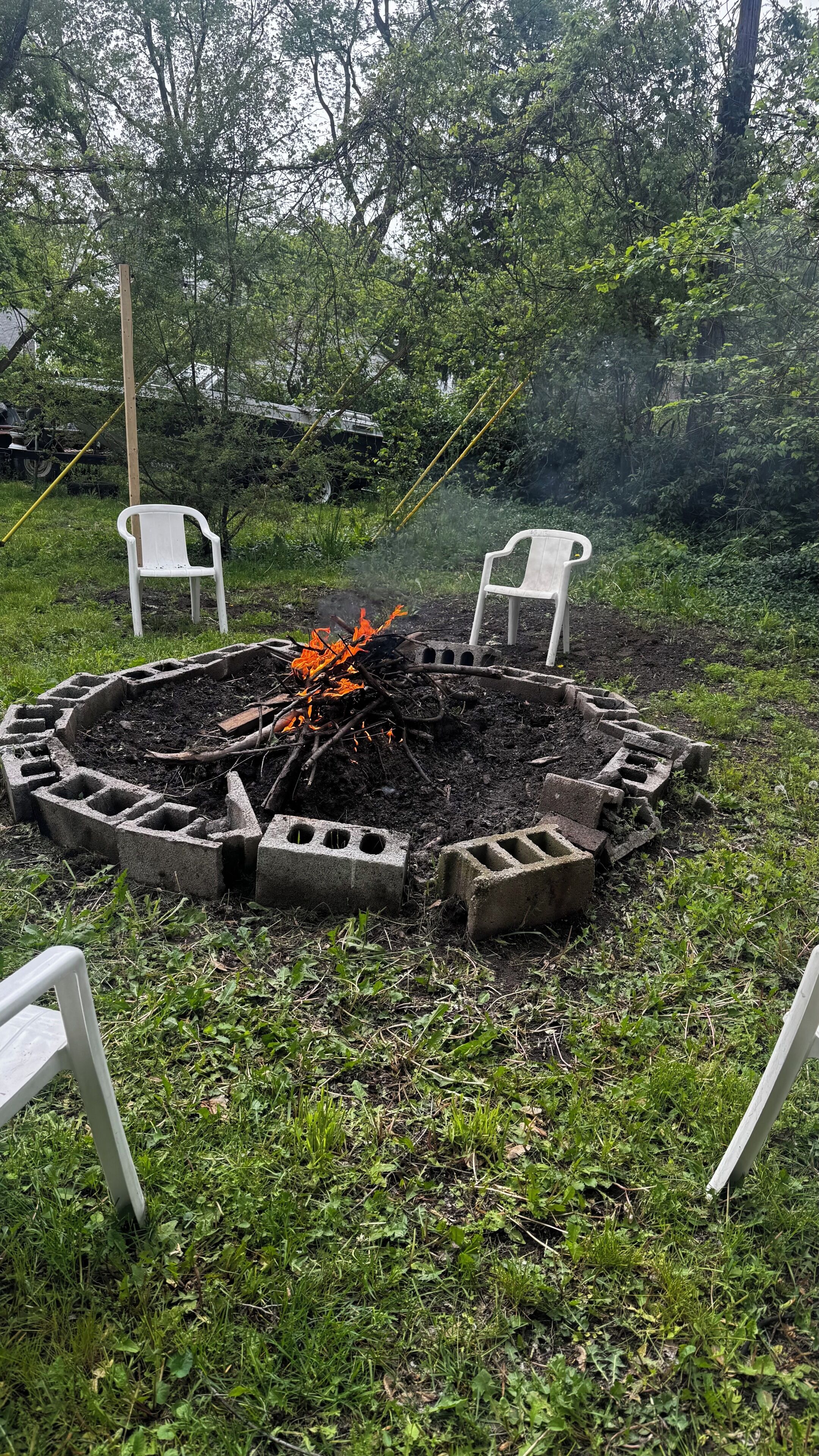 Un foyer circulaire en béton, avec deux chaises en plastique blanches de chaque côté, entouré d’herbe et d’arbres.