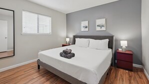 A neatly arranged bedroom with a bed, two bedside tables, a mirror, and a window with blinds. - Modern Carrollton Retreat | Pool | Sleeps 8 (Carrollton)