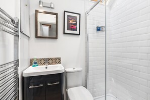 Un baño con un lavamanos blanco, un inodoro, una ducha con azulejos blancos, un espejo y un cuadro en la pared.