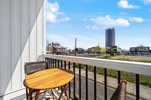 Ein Balkon mit Holztisch und Korbstuhl, mit Blick auf eine Stadtlandschaft mit Gebäuden und blauem Himmel.