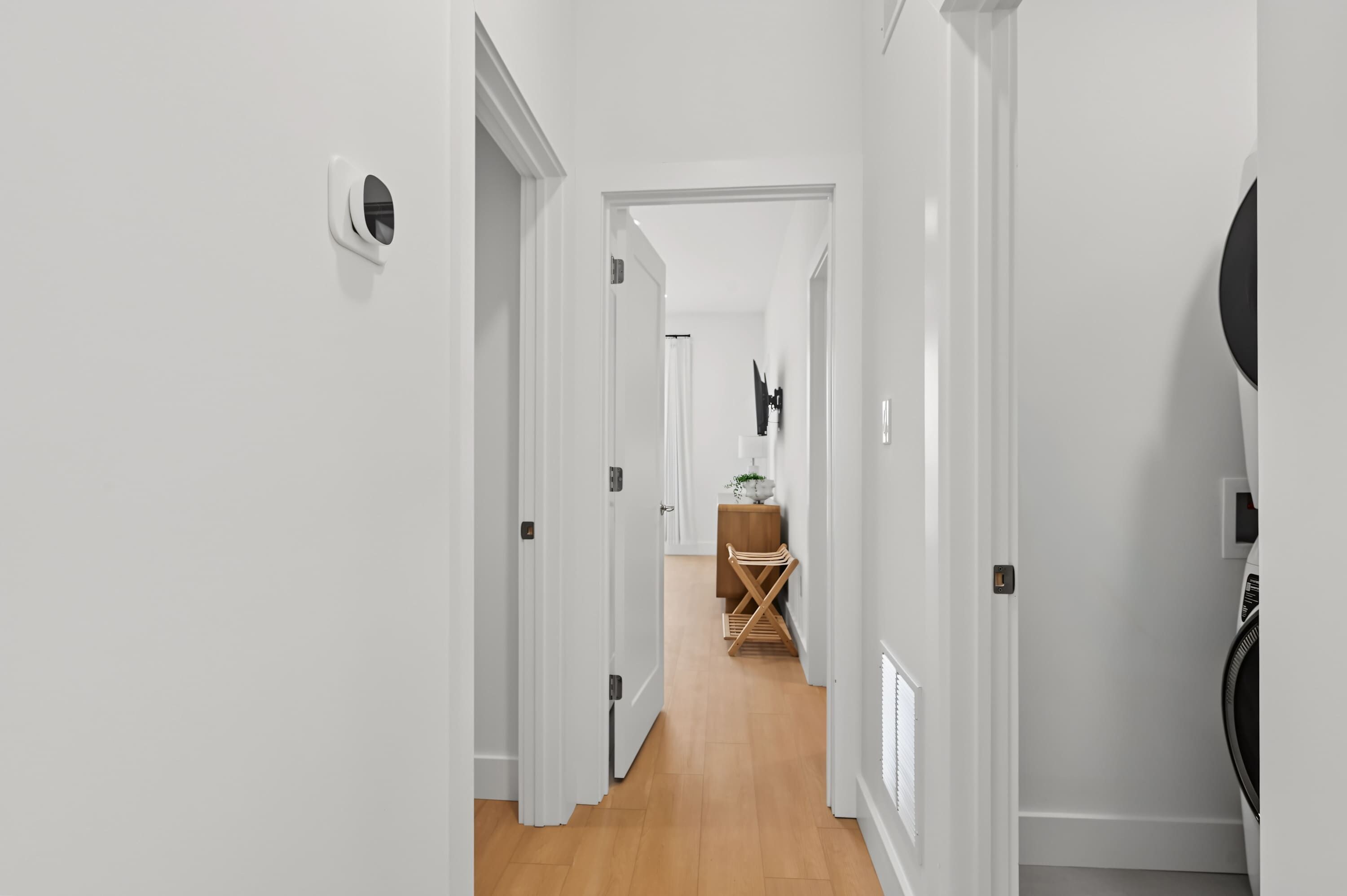 A hallway with white walls, a wooden floor, and a door leading to a room with a bed and a TV.
