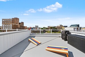 Eine Dachterrasse mit Blick auf die Stadtlandschaft, ein Grill und zwei Liegestühle.