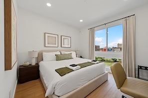 Ein Hotelzimmer mit Bett, Sessel, Nachttisch, Lampe und Fenster mit Blick auf Gebäude.