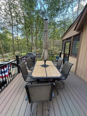An outdoor deck with a table and chairs, surrounded by a railing, and overlooking a wooded area.