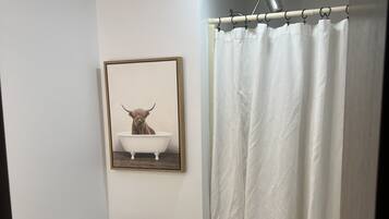 A bathroom with a toilet, a framed picture of a highland cow in a bathtub, a shower curtain, and a towel on the floor.