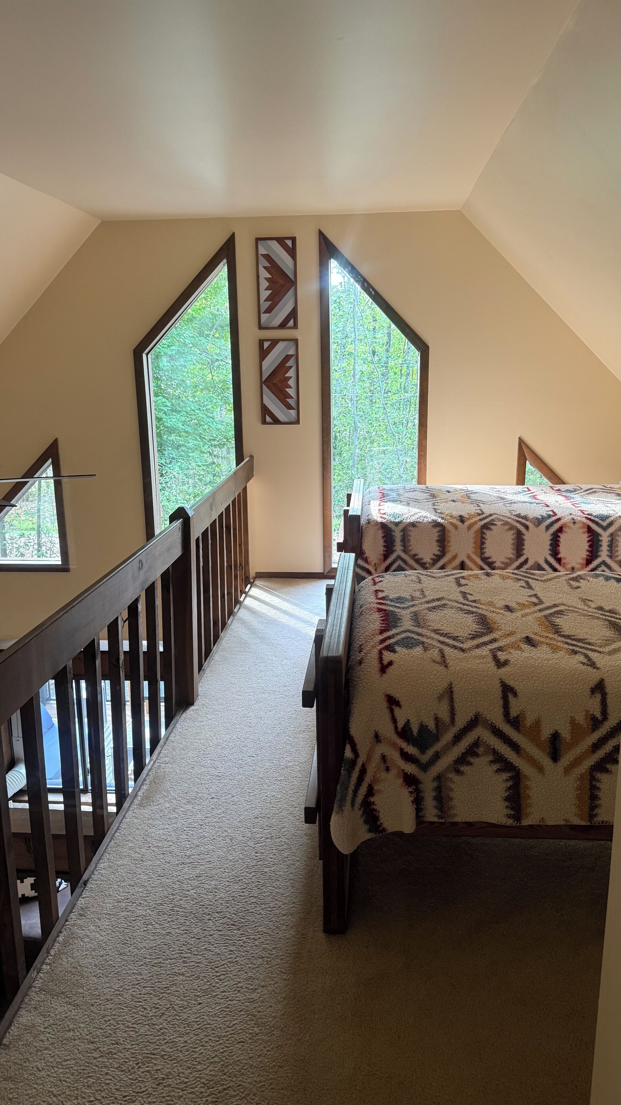 Une chambre avec un plafond en pente, un lit recouvert d’une couverture à motifs, une balustrade en bois et une vue sur la verdure grâce à de grandes fenêtres.