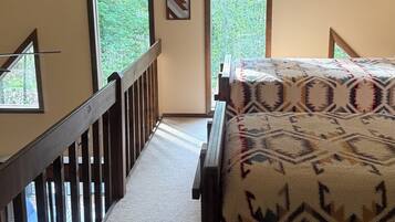 Une chambre avec un plafond en pente, un lit recouvert d’une couverture à motifs, une balustrade en bois et une vue sur la verdure grâce à de grandes fenêtres.