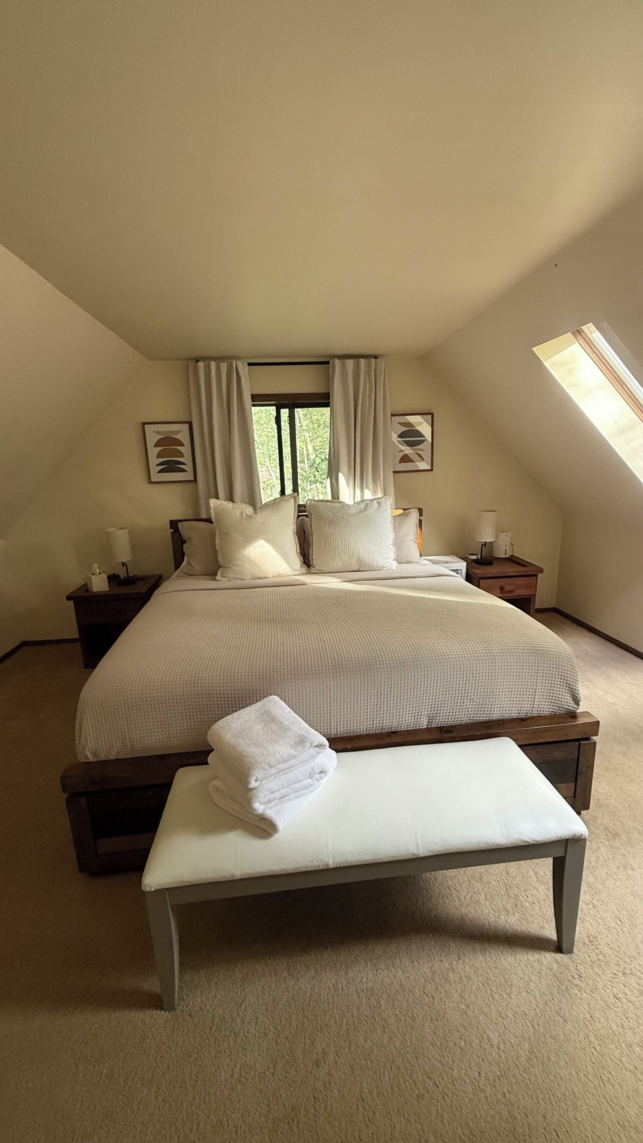 A bedroom with a large bed, a bedside table with a lamp, a bench, and a window with curtains.