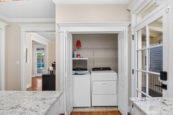 A laundry room with a washing machine and dryer, a small shelf with cleaning supplies, and a door leading to a hallway.