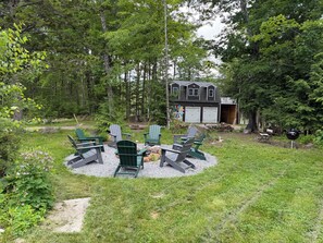 Ein kreisförmiger Sitzbereich mit Adirondack-Stühlen, einer Feuerstelle und einem Haus mit zwei Garagen im Hintergrund.
