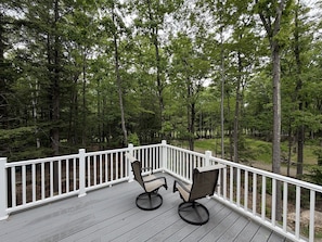 Une terrasse en bois avec des rampes blanches, deux chaises et un paysage boisé en arrière-plan.