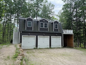 Un bâtiment à deux étages doté de trois portes de garage, d’une terrasse en bois et surplombant une zone boisée.