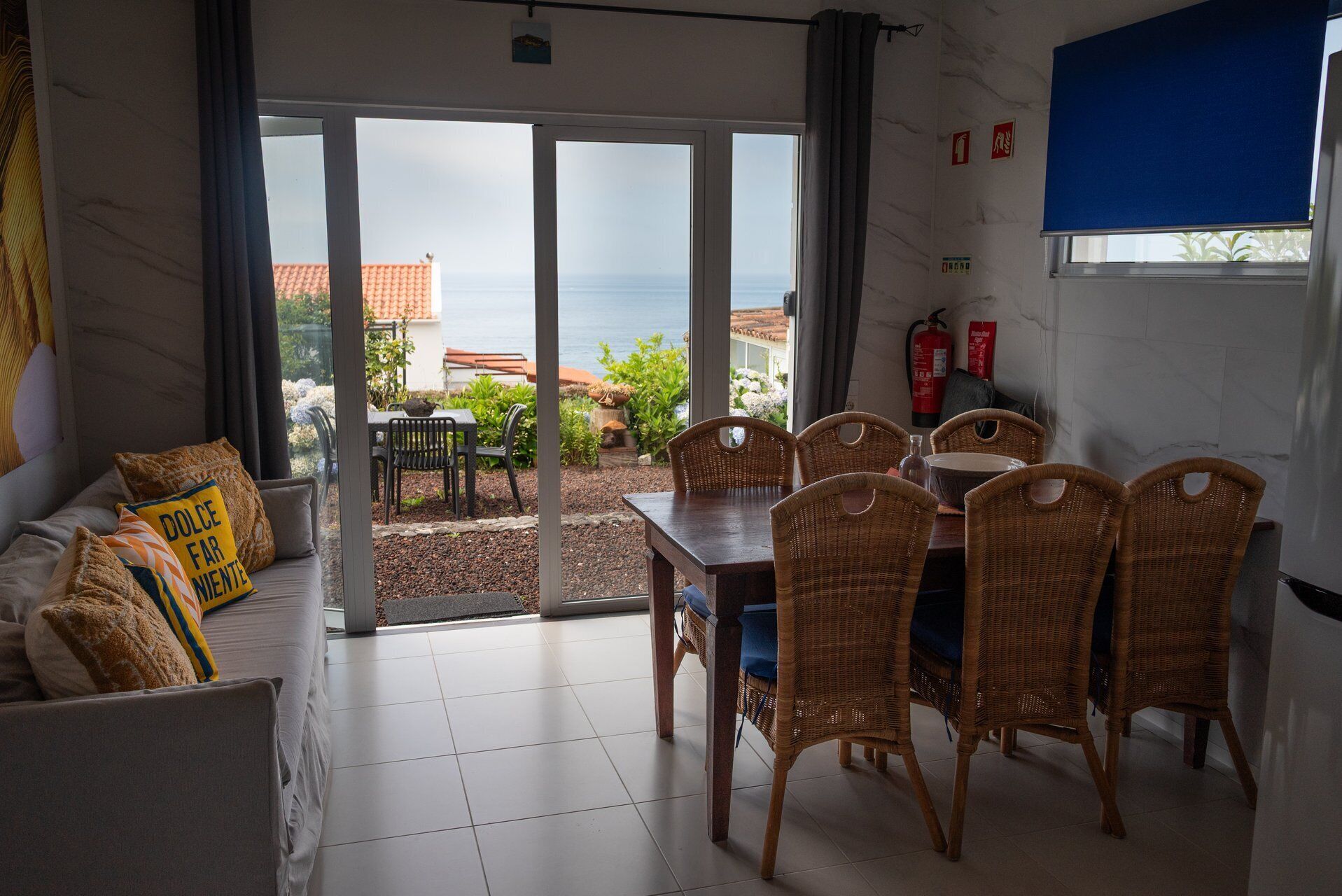 A room with a dining area, a sofa with cushions, and a view of the sea through large glass doors.