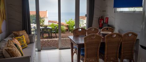 A room with a dining area, a sofa with cushions, and a view of the sea through large glass doors.