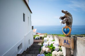 Ein schmaler Pfad führt zu einem weißen Gebäude mit Blick auf das Meer. Ein hölzernes Schild mit einem herzförmigen Emblem und den Buchstaben 'AL' ist sichtbar.