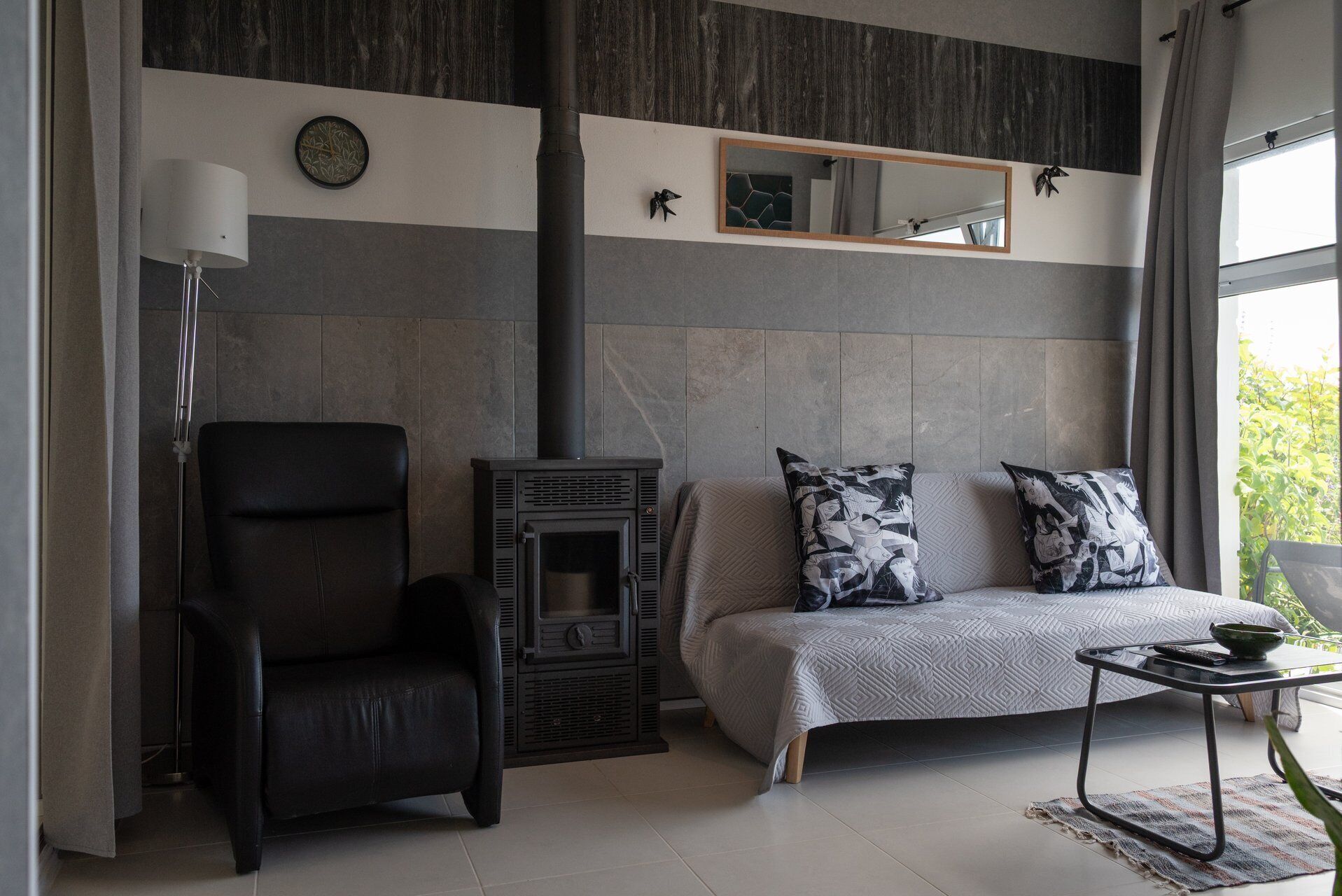 A modern living room with a black leather chair, a sofa with patterned cushions, a fireplace, and a large mirror on the wall.