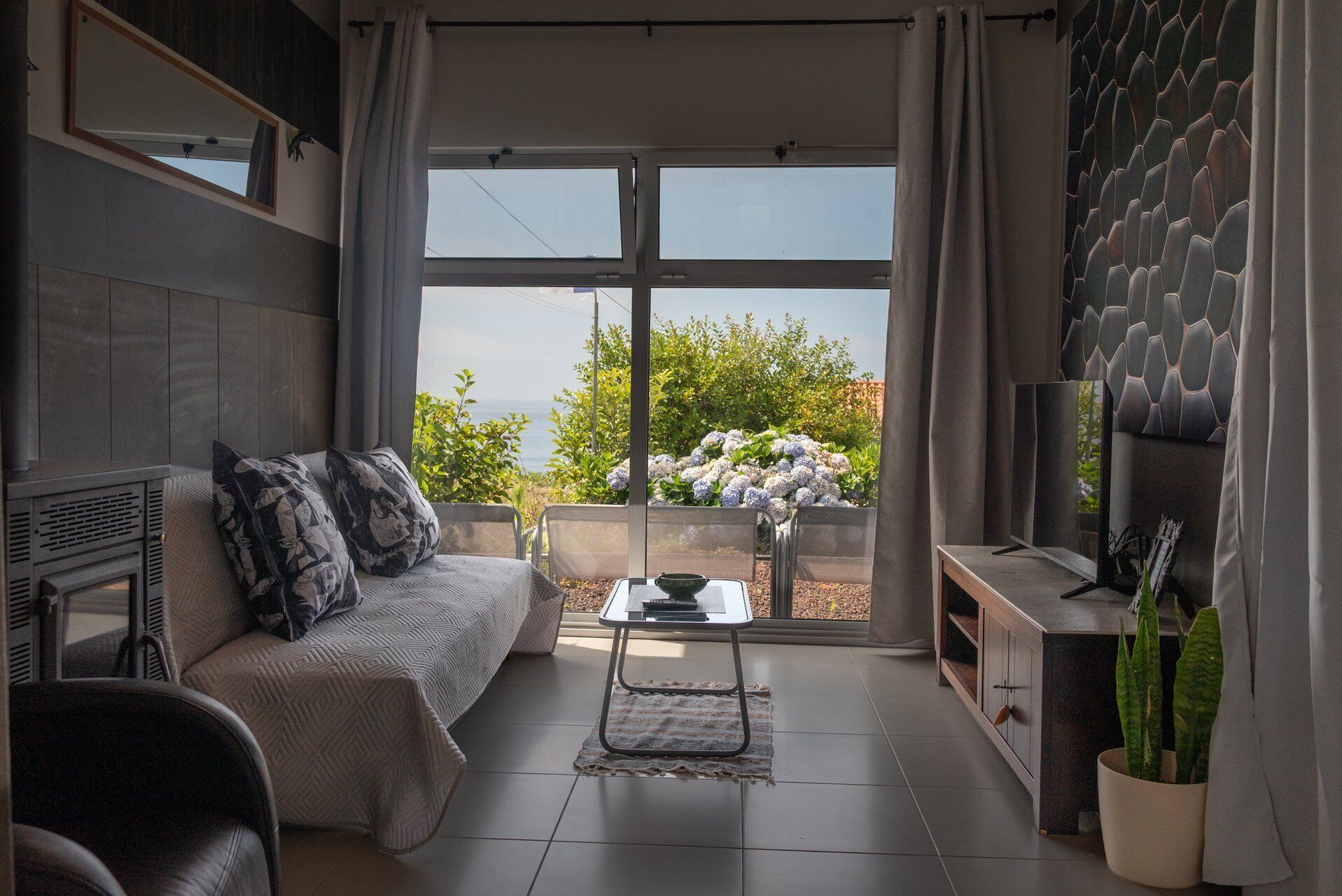 Ein modernes Wohnzimmer mit einer Couch, einem Couchtisch und einem Fernseher. Ein Balkon mit Meerblick und Pflanzen ist vorhanden.