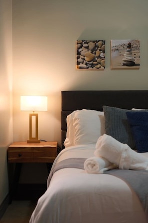 Ein ordentlich gemachtes Bett mit Kissen und einem zusammengerollten Handtuch, eine Nachttischlampe und zwei gerahmte Bilder an der Wand.