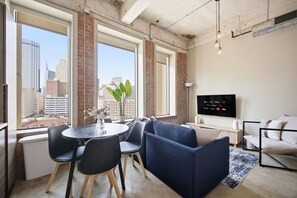 Ein modernes Wohnzimmer mit Stadtblick, ein runder Tisch mit Stühlen, ein Sofa, ein Fernseher und eine Pflanze.
