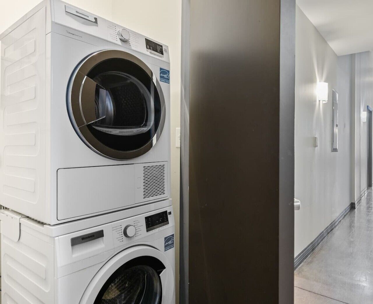 A laundry facility with stacked washers and dryers, a stainless steel door, and a hallway with a light fixture.
