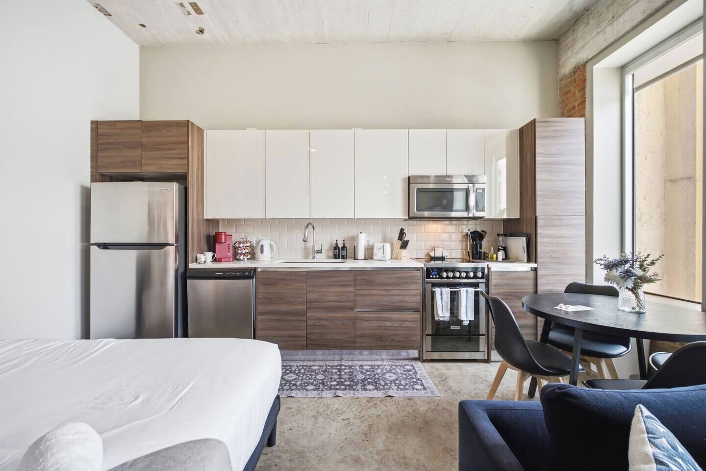 A modern kitchen with stainless steel appliances, a dining area with a round table, and a bed with a patterned rug.