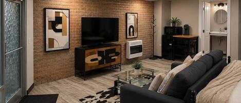 A modern living room with a brick wall, a flat-screen TV, a sofa, a coffee table, and a small kitchen area.