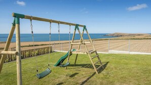 A wooden swing set with a green slide, positioned on a grassy lawn with a view of the sea and hills in the background. - Swn y Don, Gwbert - Six Bedroom Cottage, Sleeps 12 (Cardigan)