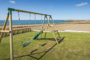 A wooden swing set with a green slide, positioned on a grassy lawn with a view of the sea and hills in the background. - Swn y Don, Gwbert - Six Bedroom Cottage, Sleeps 12 (Cardigan)