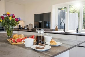 A breakfast setup with croissants, a fruit platter, a coffee maker, and a vase of flowers on a kitchen counter.