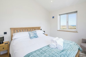 A bedroom with a wooden bed, white bedding, a blue quilted bedspread, a window with a view of the landscape, and a bedside table with a lamp and remote control.