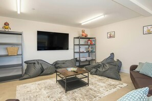 A living room with a flat-screen TV, a coffee table, grey bean bag chairs, a bookshelf, and a wall-mounted shelf. - Swn y Don, Gwbert - Six Bedroom Cottage, Sleeps 12 (Cardigan)