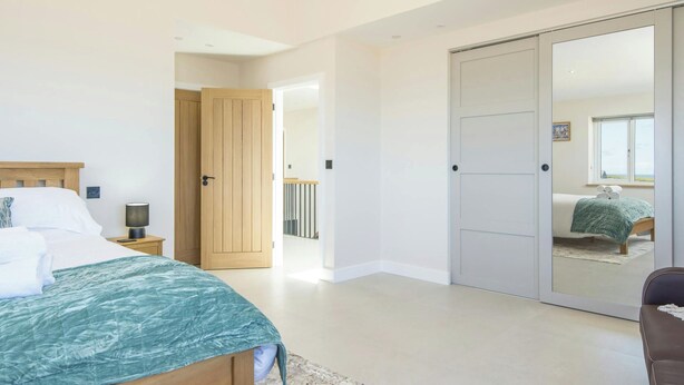 A bedroom with a bed, bedside table, lamp, and a wardrobe with a mirror. - Swn y Don, Gwbert - Six Bedroom Cottage, Sleeps 12 (Cardigan)