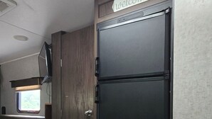 A compact kitchen area with a black refrigerator, a wooden cabinet, and a window with blinds. - Cozy Camper on Current River - Private Boat Ramp with a Dock and Views. (Doniphan)
