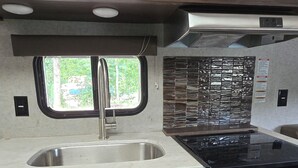 A compact kitchen with a stainless steel sink, a microwave, a stove, and wooden cabinets. - Cozy Camper on Current River - Private Boat Ramp with a Dock and Views. (Doniphan)