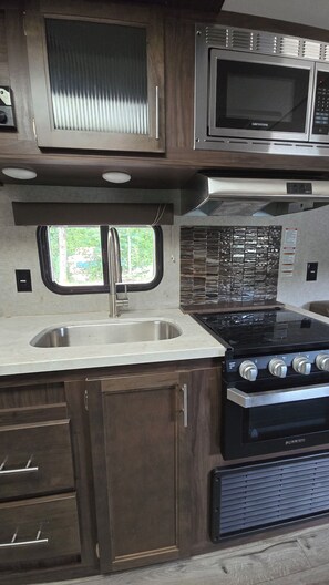 Cocina compacta con pileta de acero inoxidable, microondas, estufa y alacenas de madera.