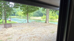 A view through a window with a gravel ground and trees, a metal structure, and a body of water. - Cozy Camper on Current River - Private Boat Ramp with a Dock and Views. (Doniphan)