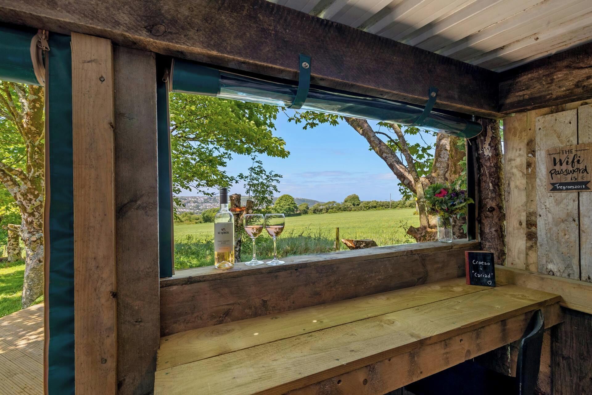 A rustic wooden window with a view of a green landscape, a bottle, and a glass on a wooden ledge.
