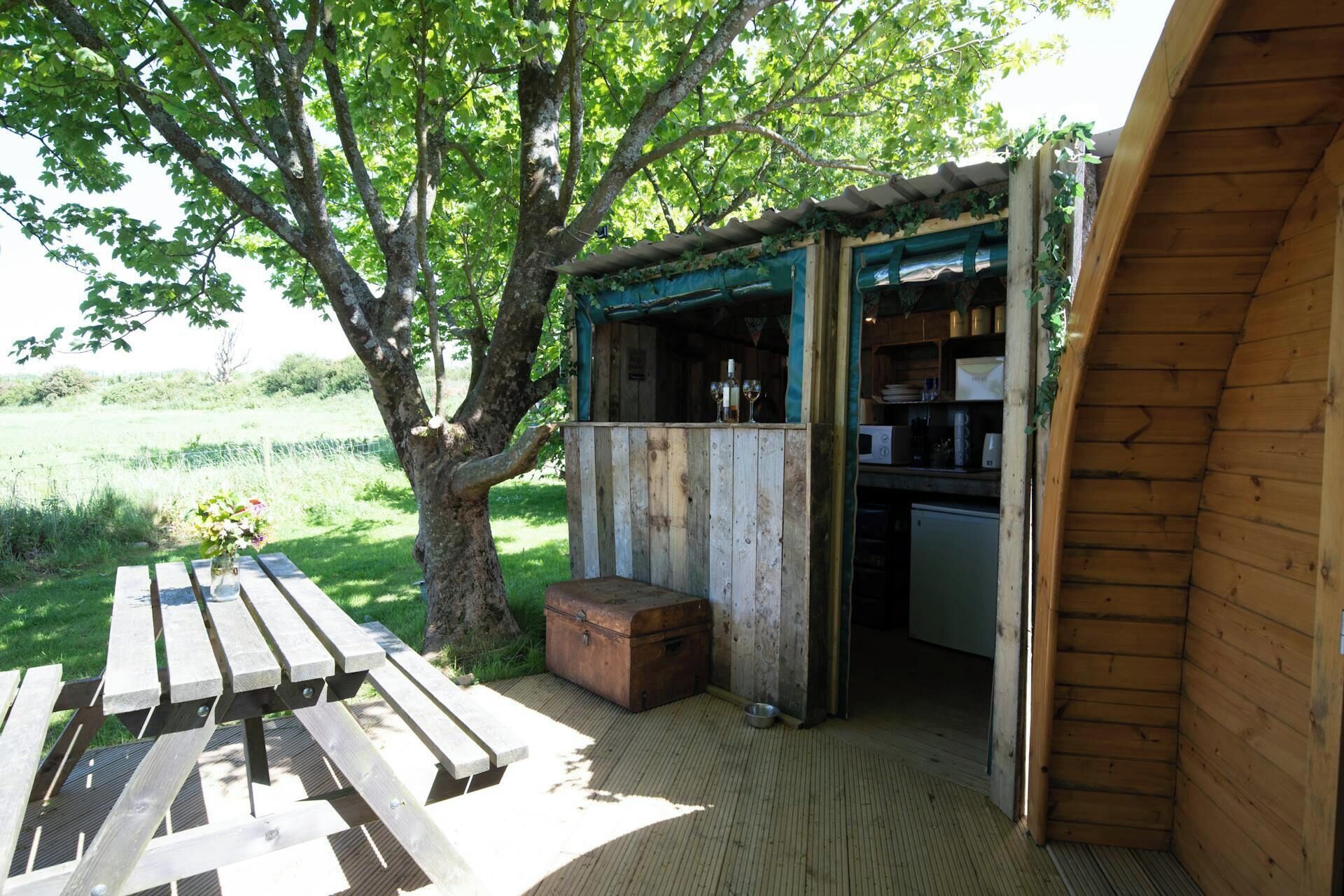 Une structure rustique en bois, avec un banc et une table, un petit coin cuisine et un arbre en arrière-plan.