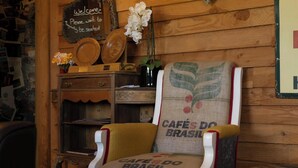 A wooden interior with a chair featuring a 'CAFE DO BRASIL' cushion, a round table with a 'WELCOME' sign, and a wall-mounted shelf with decorative plates.