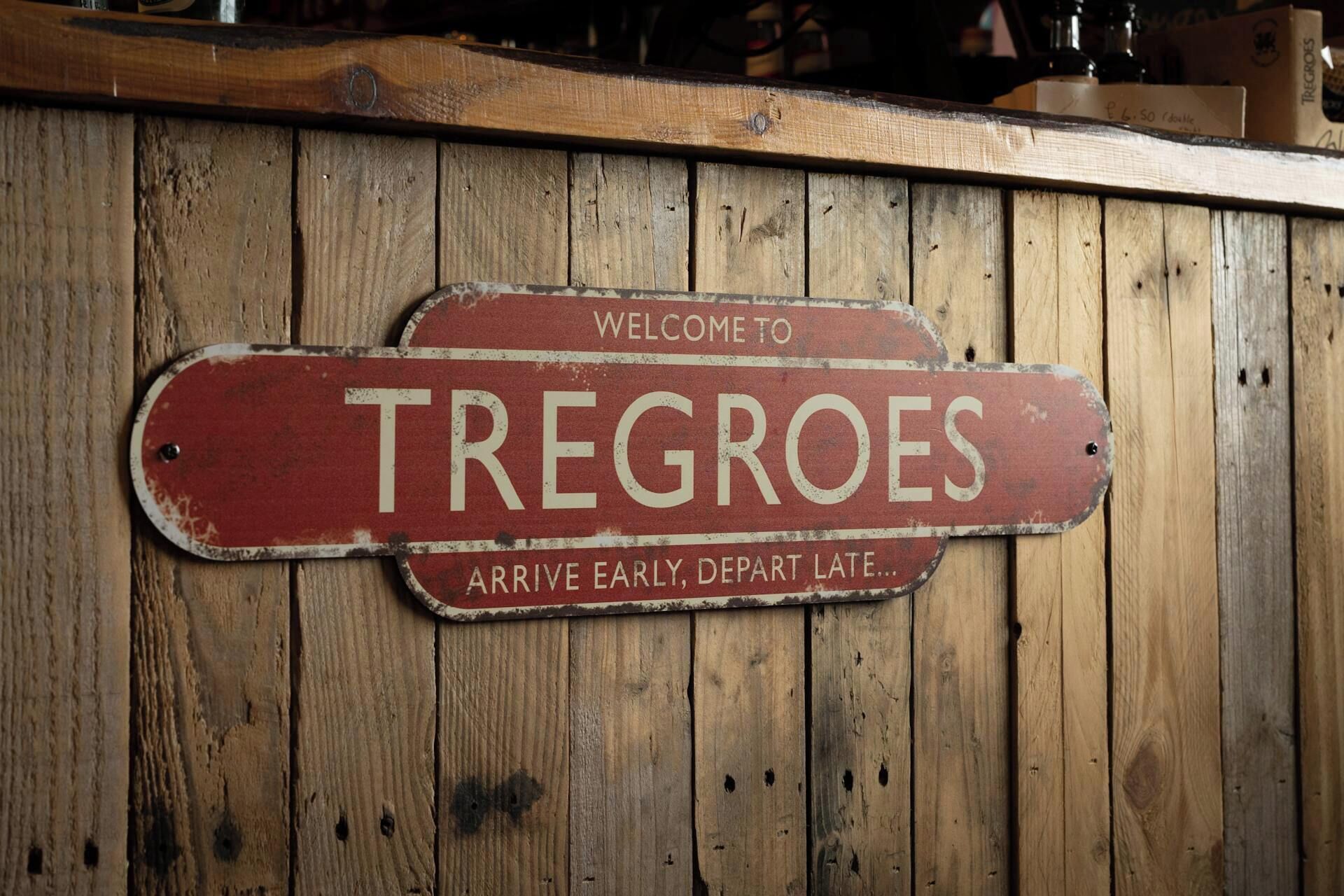 Eine Holzwand mit einem roten Schild, das 'WELCOME TO TREGROES' zeigt und dazu rät, 'früh anzukommen und spät abzureisen'.