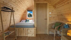 A wooden cabin room with a loft bed, a window with a view, a door, and a wicker chair.