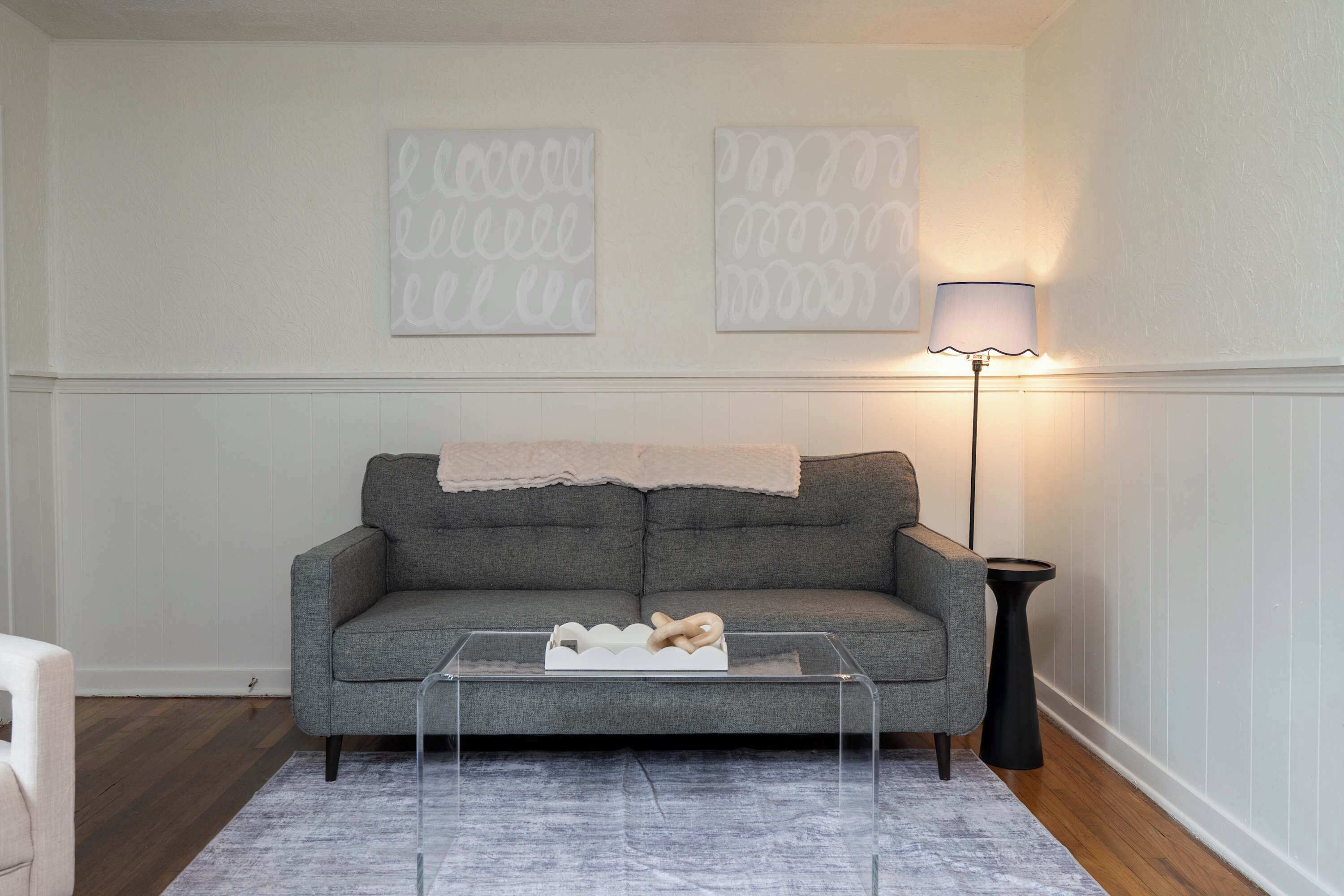 A living room with a grey sofa, a glass coffee table, a lamp, and two wall art pieces.