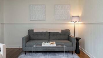 A living room with a grey sofa, a glass coffee table, a lamp, and two wall art pieces.