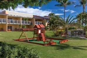 A playground with a slide and climbing structure, surrounded by a well-maintained lawn and palm trees. - Co2bedroom condo with AC in fantastic Kailua-Kona (Kailua-Kona)