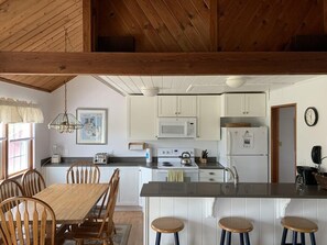 A kitchen with white cabinets, a microwave, a stove, and a refrigerator. There is a dining table with chairs and a bar stool. - Charming Nantucket Cottage w/ Private Beach Access - 50%! (Nantucket)