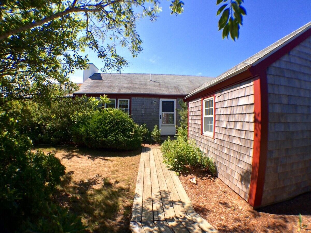 Une maison dotée d’une terrasse en bois menant à la porte d’entrée, le tout entouré de verdure.