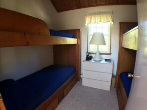 Quarto compacto com beliches, uma cômoda pequena e janela iluminada por um abajur.