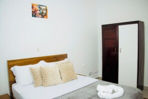 A bedroom with a wooden headboard, a bed with white bedding and beige pillows, a gray blanket, and a white towel on the floor. - 3K Residences Yaoundé Odza - Premium 2-Bedroom Apartment - D3 (Yaoundé)