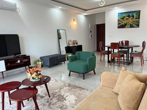 A living room with a sofa, a green armchair, a coffee table, a TV, and a dining area with chairs and a table. - 3K Residences Yaoundé Odza - Premium 2-Bedroom Apartment - D3 (Yaoundé)